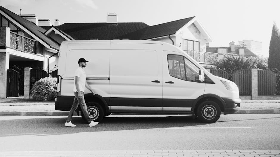 A man wearing a white t-shirt and jeans is loading cardboard boxes onto a white van parked on the street in a residential area during daytime. The open rear doors of the van reveal a space prepared fo