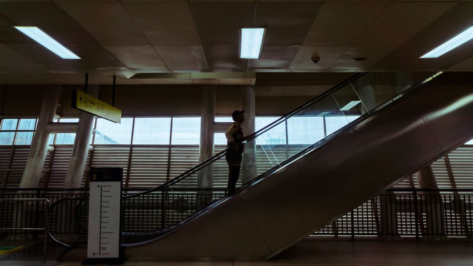 A man with gray hair, seen from behind, standing on an escalator inside a building, holding onto the handrail with his right hand. He is wearing a dark jacket and light-colored trousers. The ceiling a