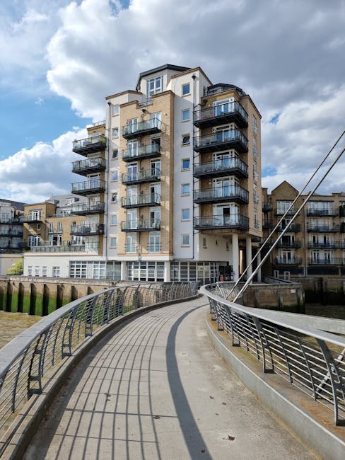 A multi-storey residential building with multiple balconies featuring glass railings, situated adjacent to a waterfront in an urban area. The building has a modern design with beige and white exterior walls, and several windows visible on each floor. A curved concrete walkway with metal railings runs along the water's edge, leading towards the building, with shadows cast by the railings. The sky is partly cloudy with patches of blue. This scene exemplifies a typical setting for house removals and relocation services provided by Lewisham Man and Van, where careful planning is needed to transport furniture and boxes across similar properties and scenic urban environments.