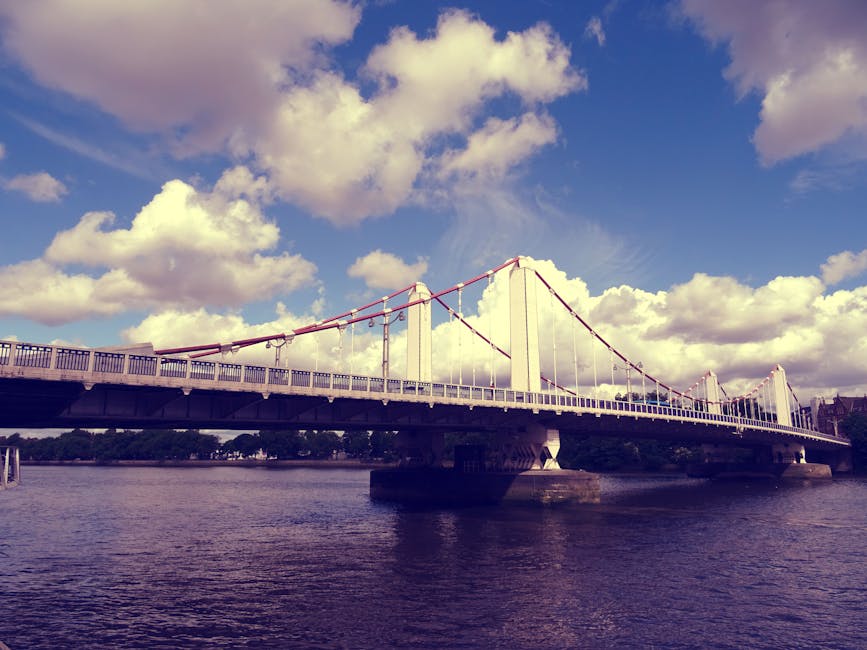 Photograph of a large modern bridge spanning a wide river, with tall white towers supporting red suspension cables. The bridge’s deck is lined with safety railings, and the sky above features scattered white clouds against a blue background. The river below reflects the bridge structure, and there are no visible vehicles or pedestrians on the bridge. The image is taken during daylight, highlighting the structural elements and open sky. This scene is relevant for house removals and moving services by Lewisham Man and Van, illustrating the importance of efficient transport infrastructure in home relocation and furniture transport processes.