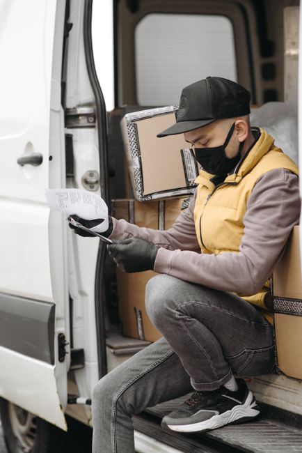 A male movers from Lewisham Man and Van wearing a black face mask, white t-shirt, and blue work overalls with black straps is holding a cardboard box filled with packing materials. He is standing outd