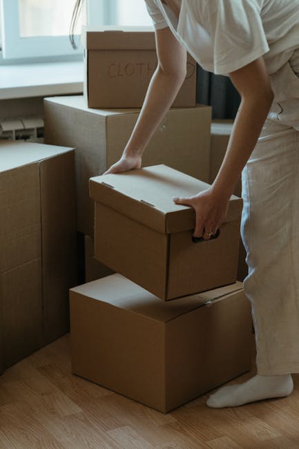 A male mover with a beard and curly hair, wearing a navy blue t-shirt with a logo on the chest and dark trousers, is inside a bright, spacious room with white walls and a large arched window in the ba