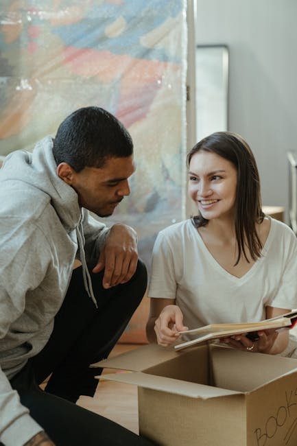 A man and a woman are engaged in a home relocation process, with the man kneeling on the floor and the woman sitting inside a room. The woman, holding a book or folder, is smiling and appears to be di