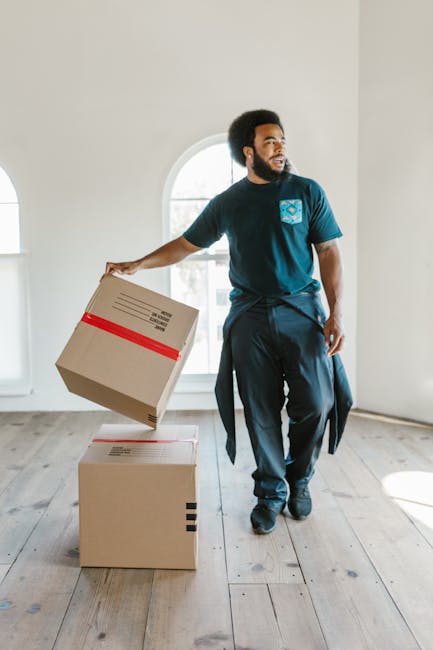 A young woman with curly black hair, wearing a white t-shirt, light blue jeans, and white sneakers, is sitting cross-legged on a wooden floor surrounded by packed cardboard boxes of various sizes, som