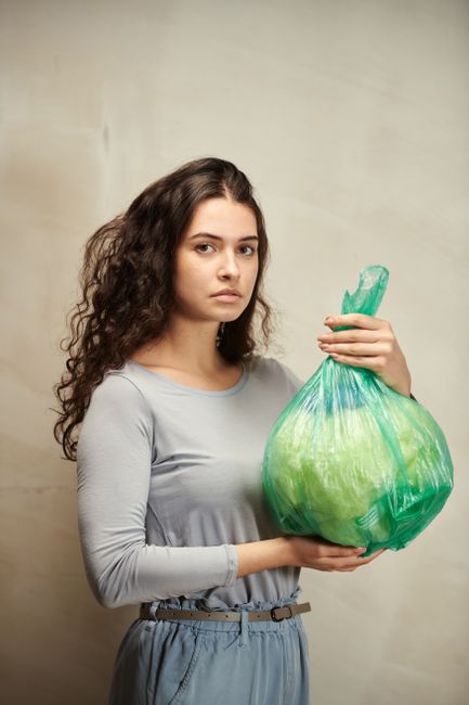 A young woman with long, curly brown hair and a serious expression is standing indoors, holding a large green plastic bag filled with items, presumably for a house move. She is wearing a light grey lo