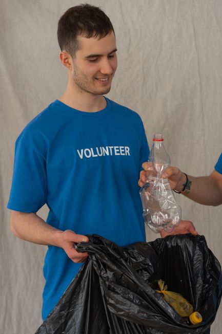 A young woman with long, curly brown hair and a serious expression is standing indoors, holding a large green plastic bag filled with items, presumably for a house move. She is wearing a light grey lo
